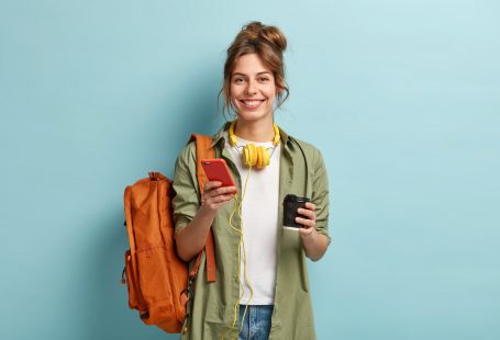 Studio shot of glad female student has coffee break after lectures, listens audio book in headphones, enjoys record from website, uses mobile phone for chatting online, has rucksack on back.